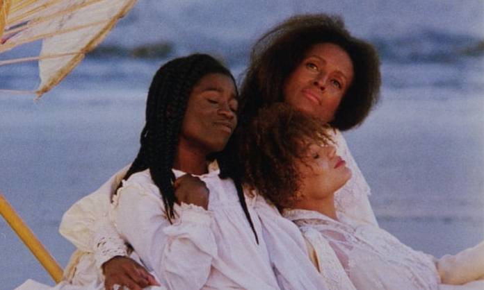 Film still from Daughters of the Dust; three Black women sit dressed in white with the ocean behind them