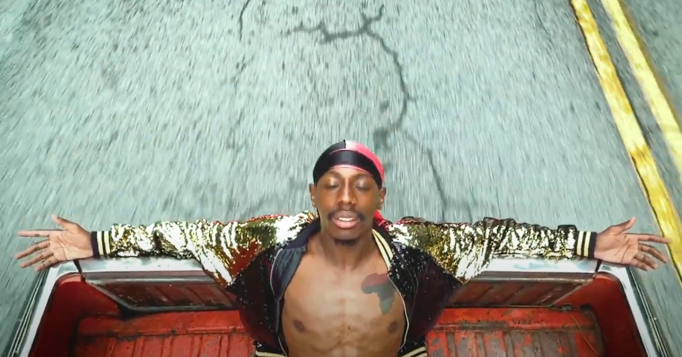 A Black man sits in the back of a truck with his arms outspread and eyes closed, wearing a sequined gold jacket that parts to reveal a tattoo of the African continent on his chest