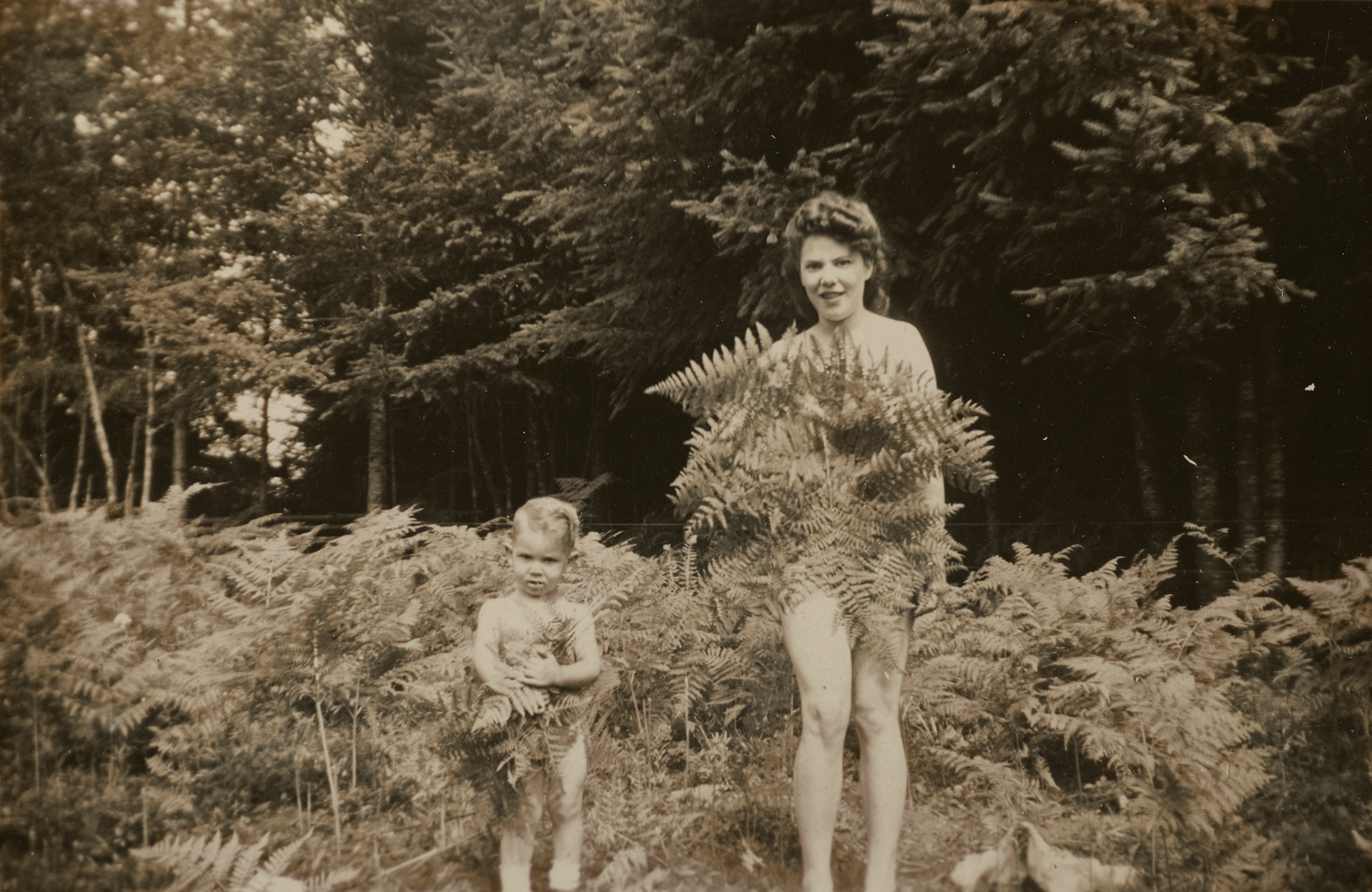 Snapshot of a light-skinned woman and young child standing naked in a bed of ferns at the edge of a forest. The woman smiles, both covering themselves with fern fronds.