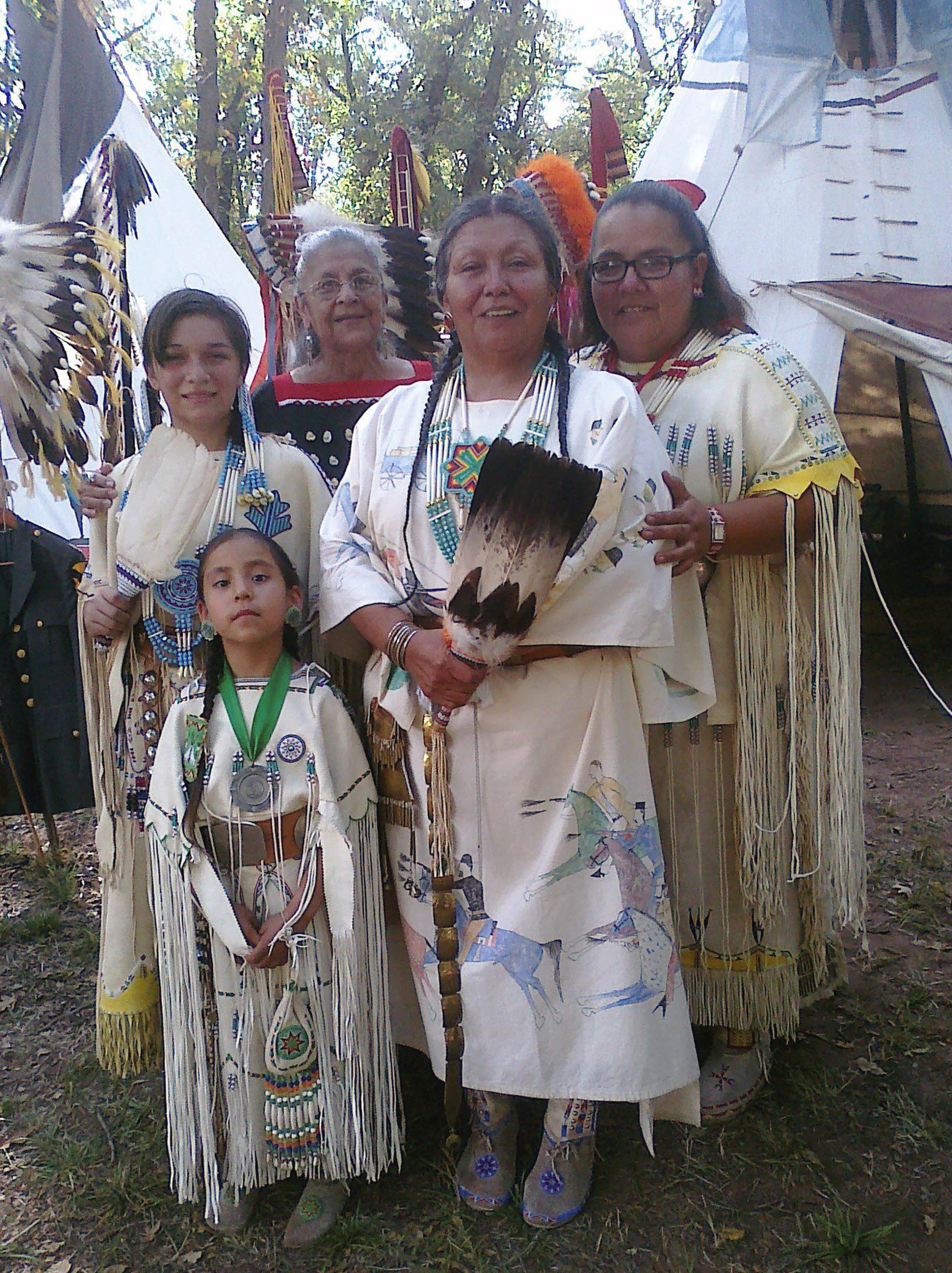 Five Indigenous women and girls stand between two tepees. They wear decorated buckskin dresses and hold feather fans. They look at the viewer, and several of them smile.