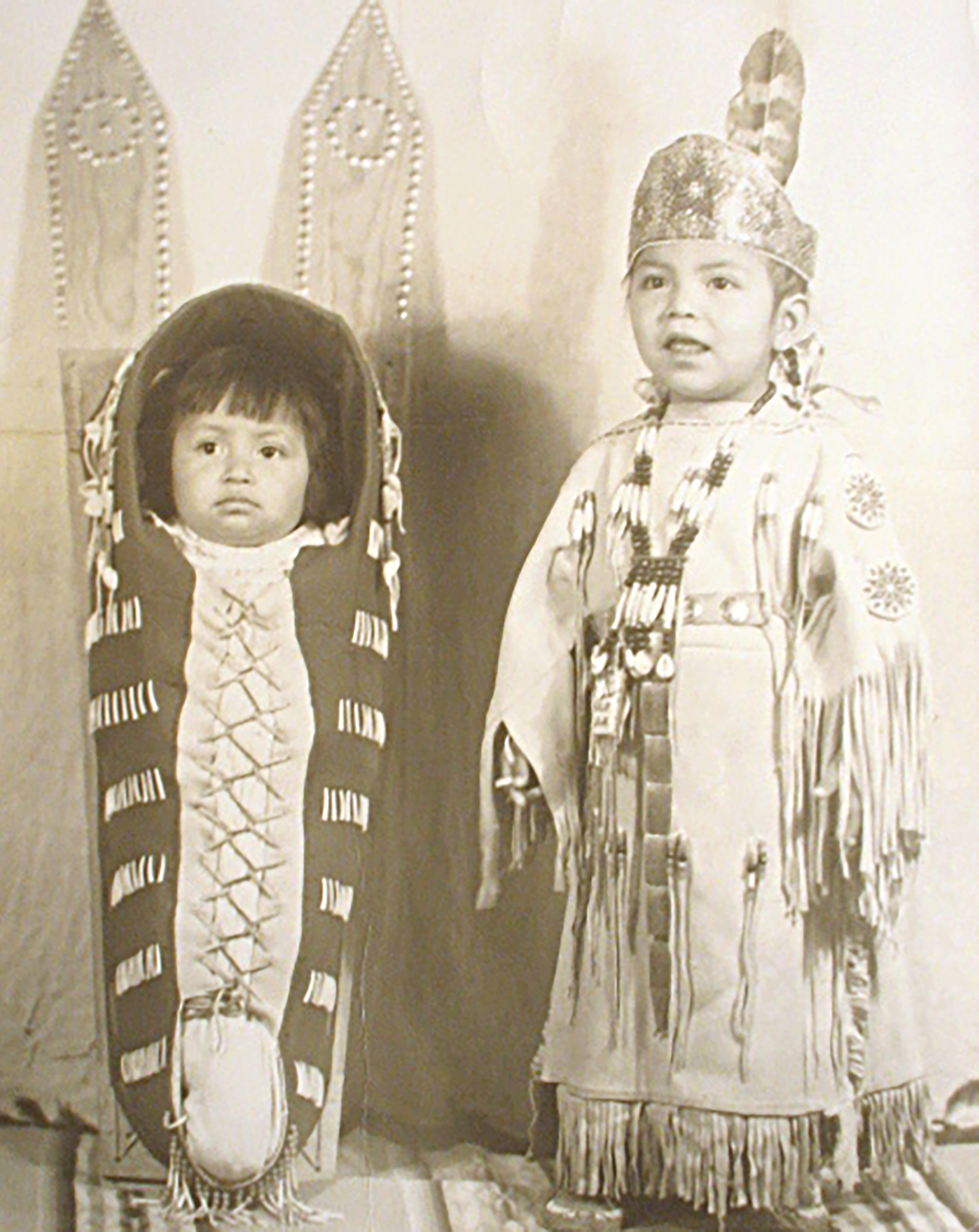 Black and white photo of two Indigenous children. A baby is laced into a carrier at left, and a toddler in a beaded buckskin dress stands at right.