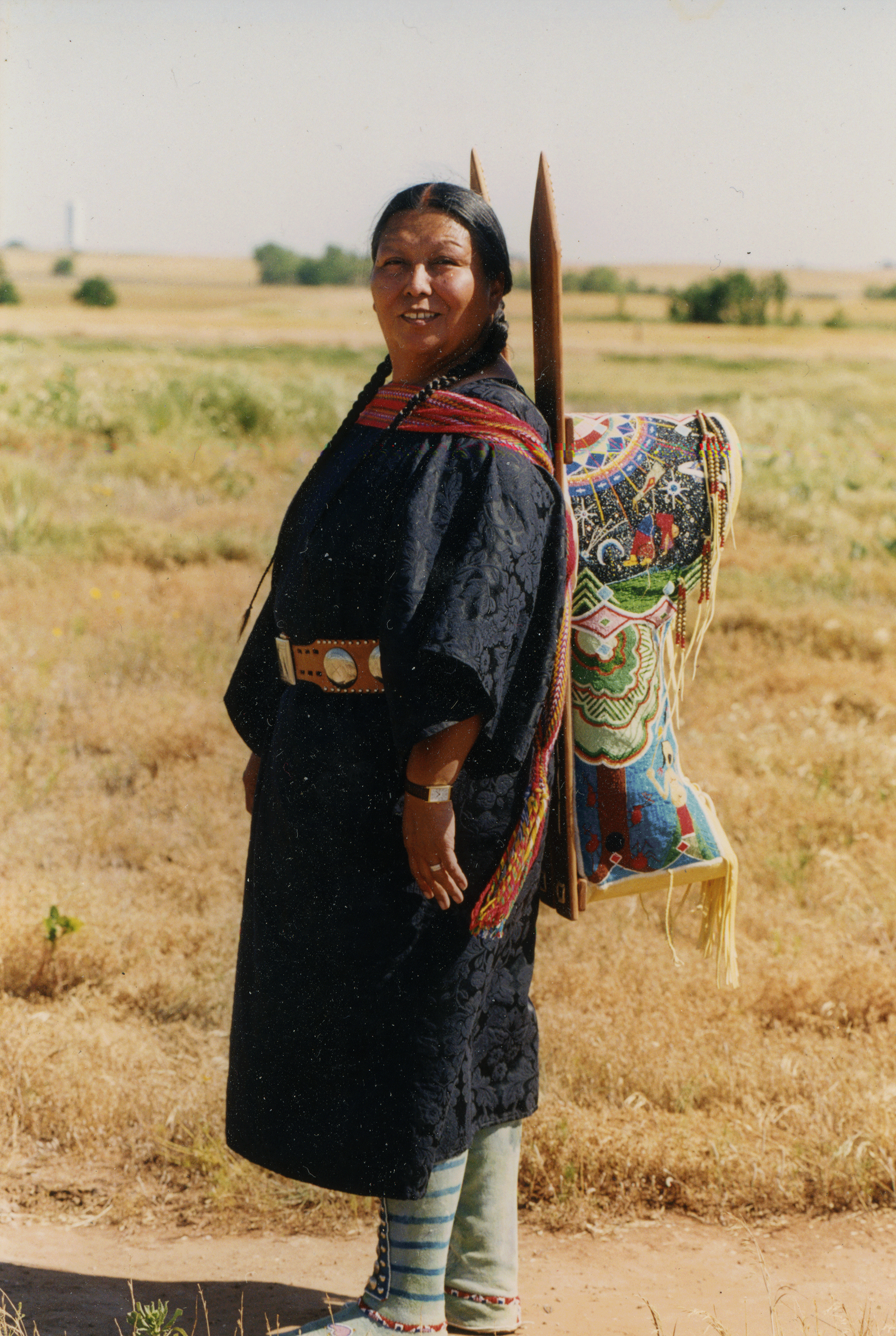 Brown-skinned woman wearing a colorful baby carrier strapped to her back. Her hair is braided and she wears a dark dress and striped socks.