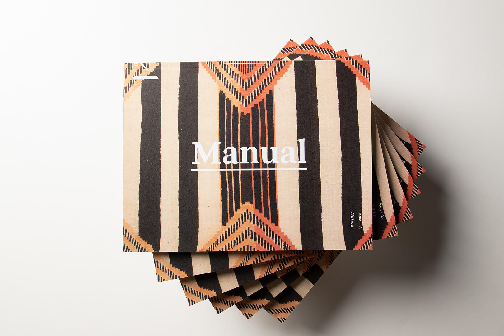 A stack of magazines arranged in a spiral formation photographed from above. The magazine cover includes a black and tan striped textile with orange decorative motifs and the word Manual in white across the top. 