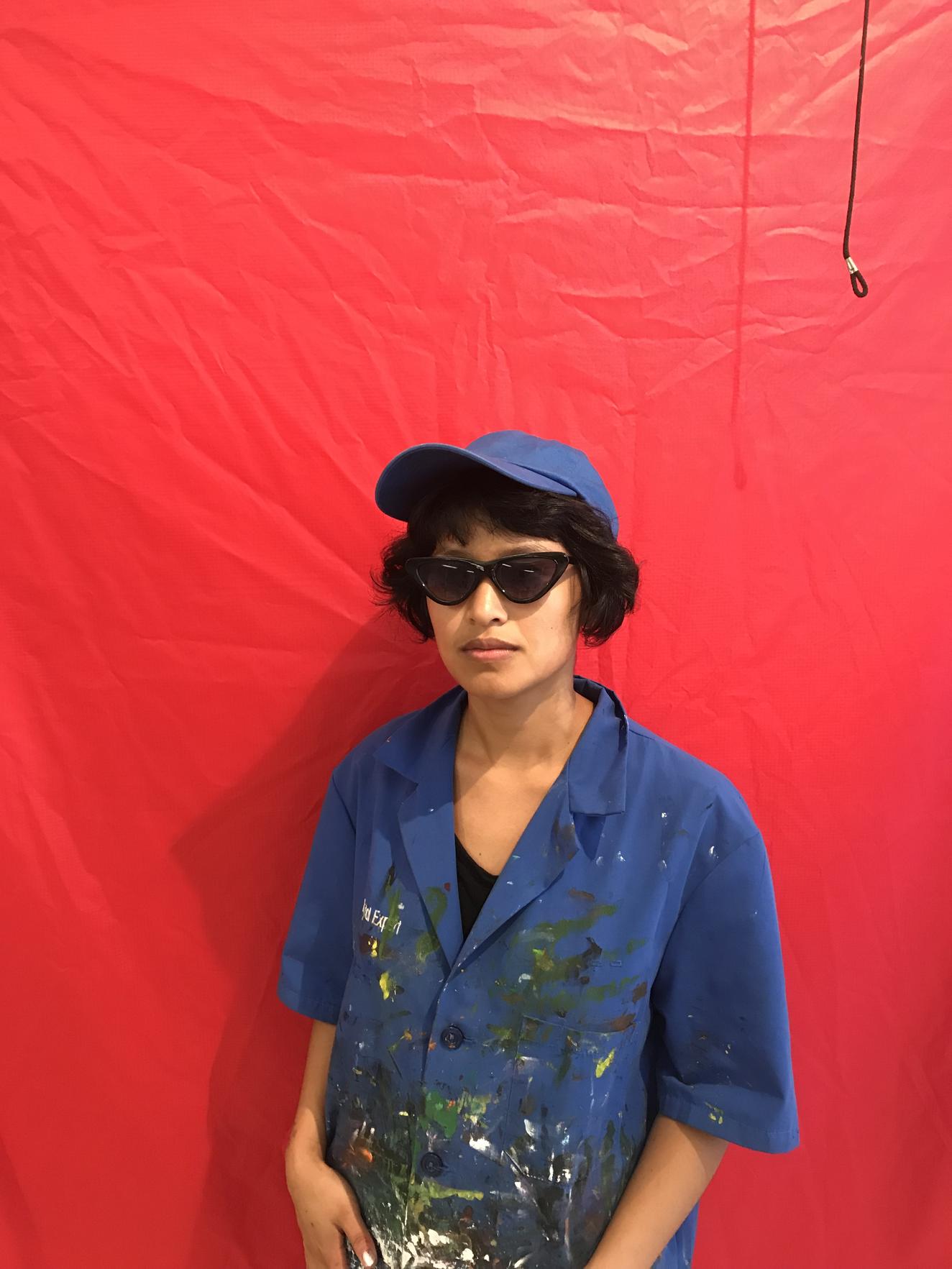 Image of 2019 Artist Fellow Dana Heng. Woman with blue hat and shirt with paint splotches, wearing red sunglasses in front of a red background.