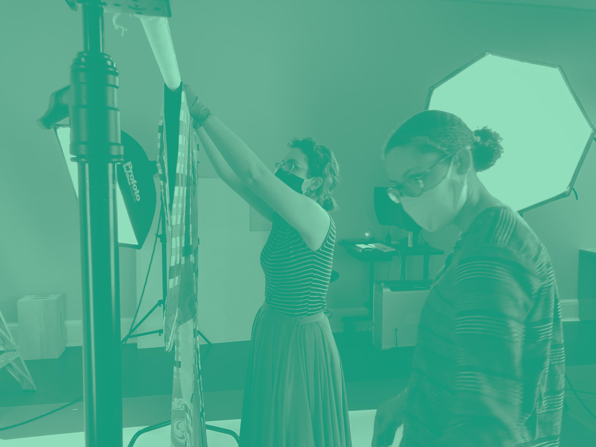 Two masked people set up photography for a textile. A large lamp shines behind them. A green filter has been applied to the photo.