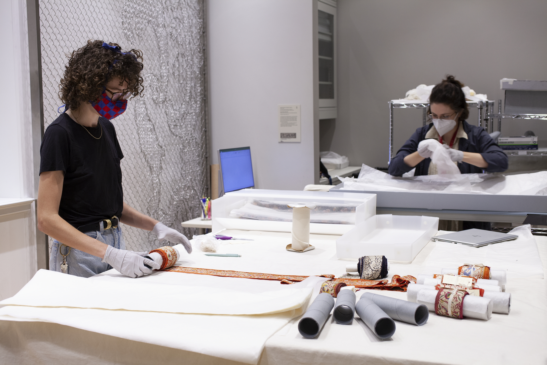 A masked museum worker stands at a table, materials laid out before them. A second person works sitting at a table in the background