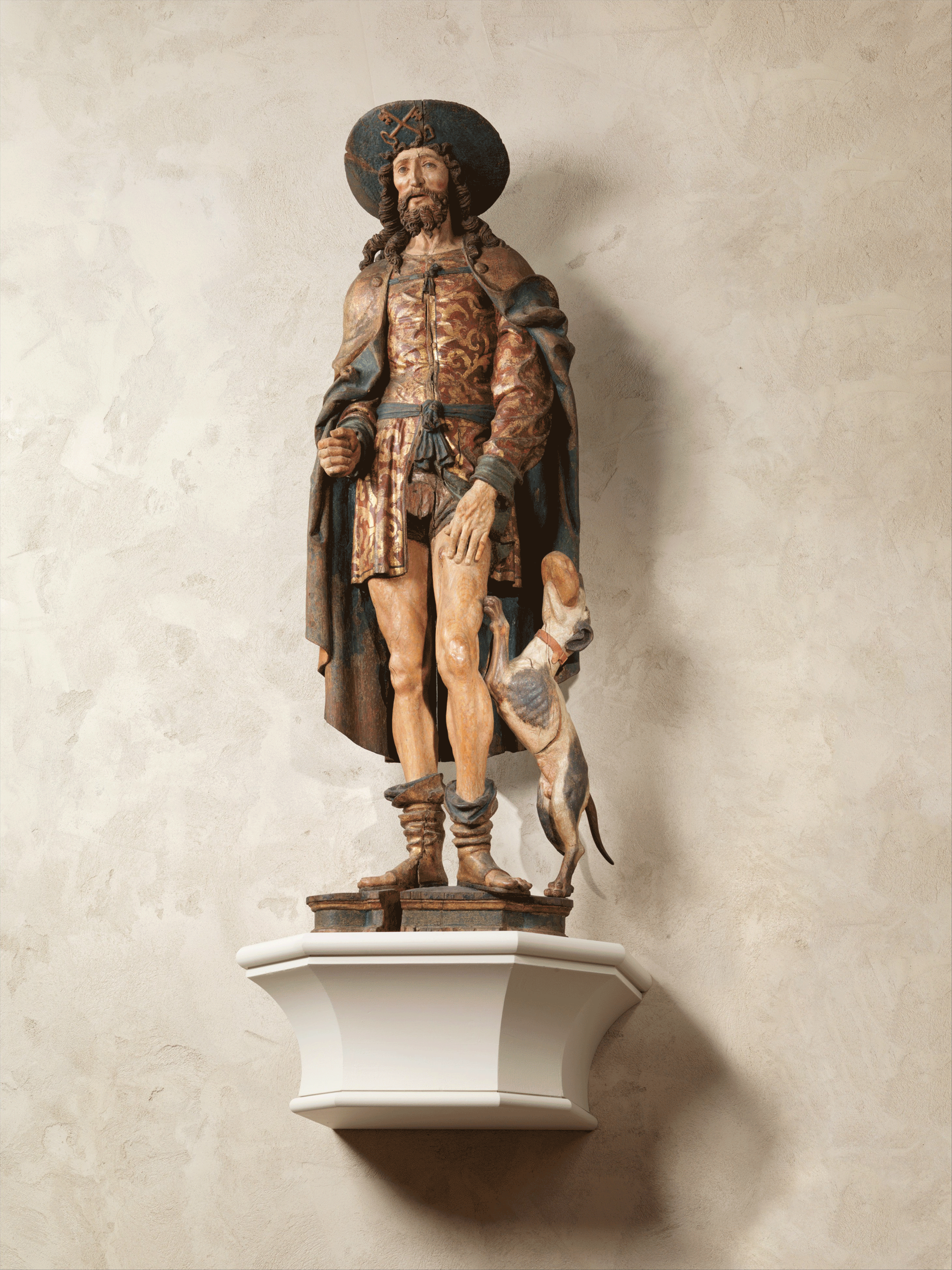 Painted sculpture of a bearded man in an elaborate short tunic and cloak. His left hand rests on his bare thigh. A dog stands on his back legs beside him.