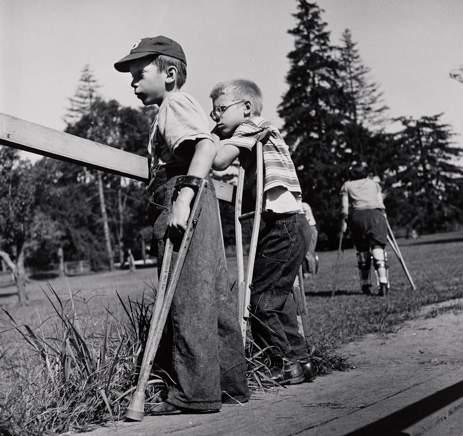 Two light-skinned boys stand outside in profile, supported by crutches, watching something out of frame. Behind them, another person in leg braces walks away using crutches. 