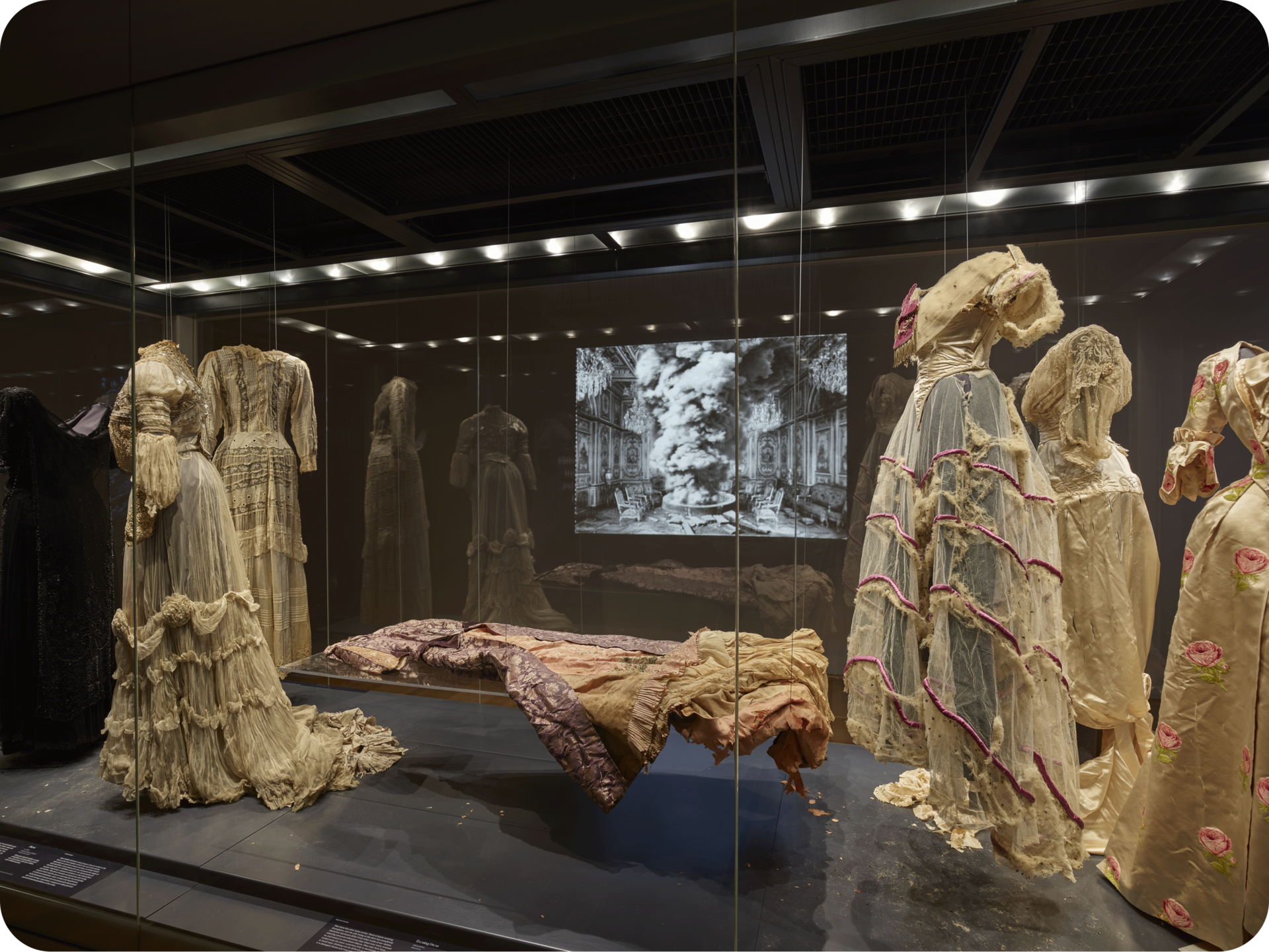 View of several ornate cream-colored dresses on mannequins encased in glass. A table in the center holds another dress. In the background is a projection of a grand ornate space. 