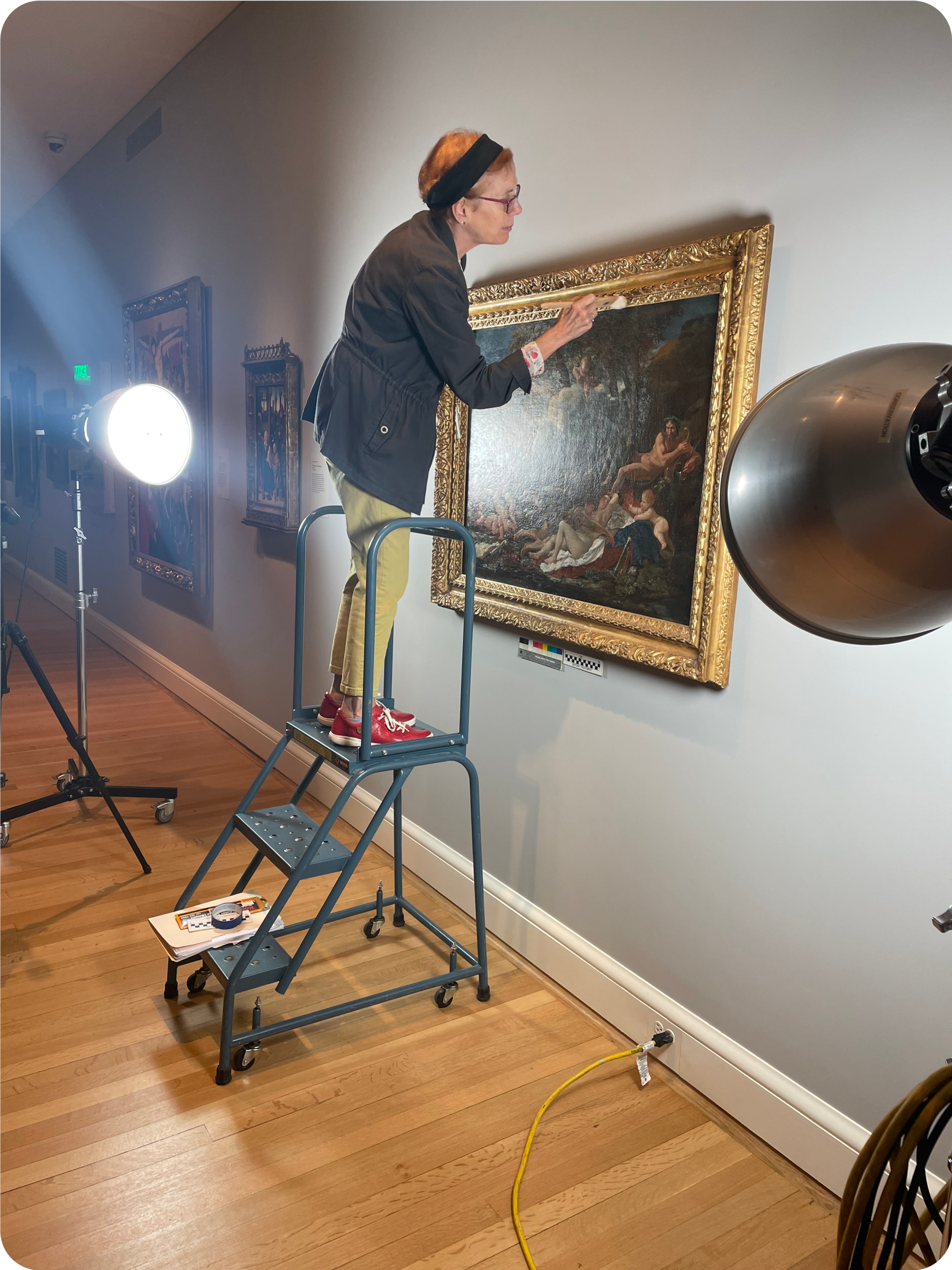 Person standing on the ladder holding a paintbrush to a painting’s ornate golden frame. The painting is hung on a gray wall and a bright lamp shines onto the painting. 