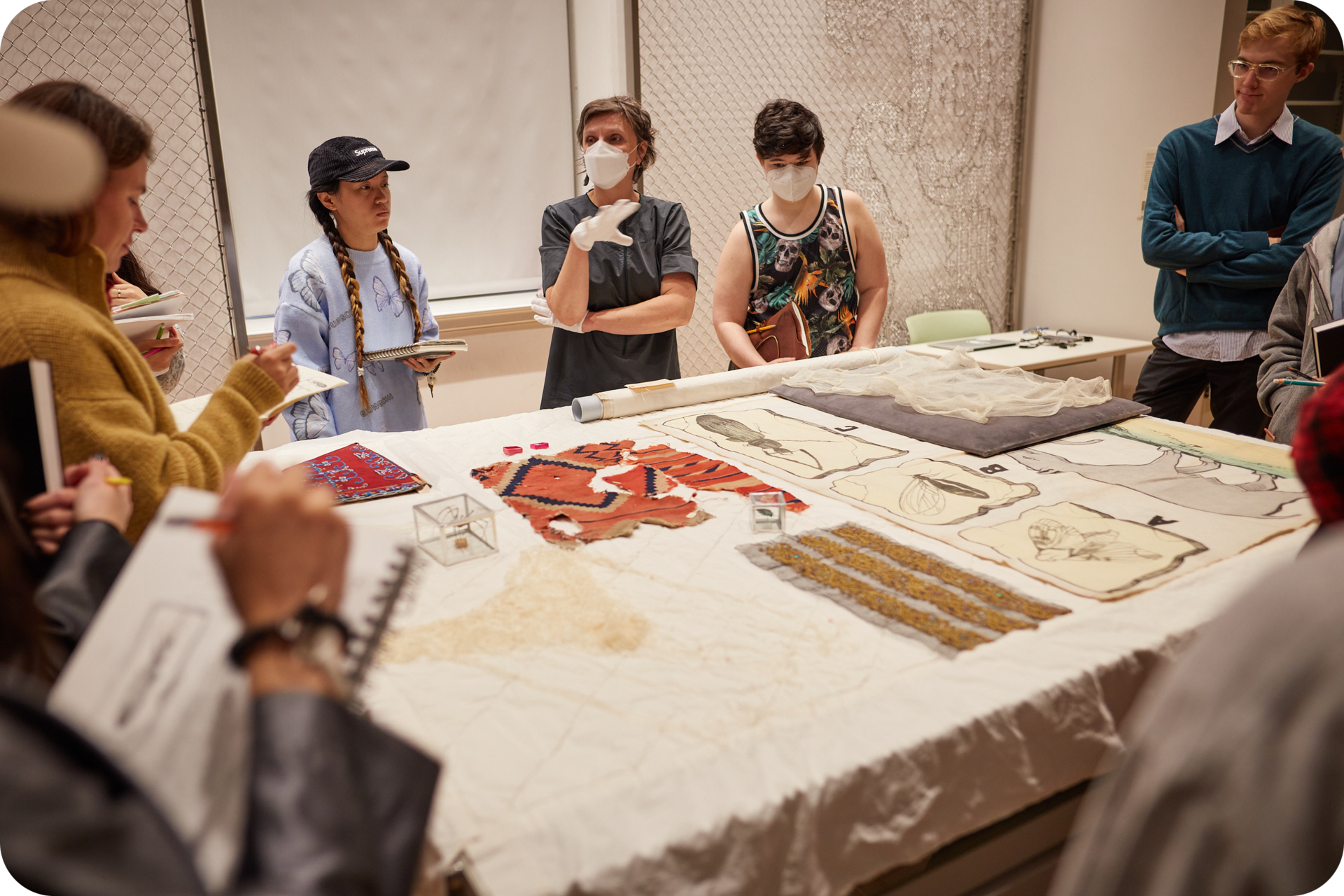 Several people standing around a table displaying various textiles. A person in the center wearing gloves gestures towards another person, while others stare at the textiles on the table. 