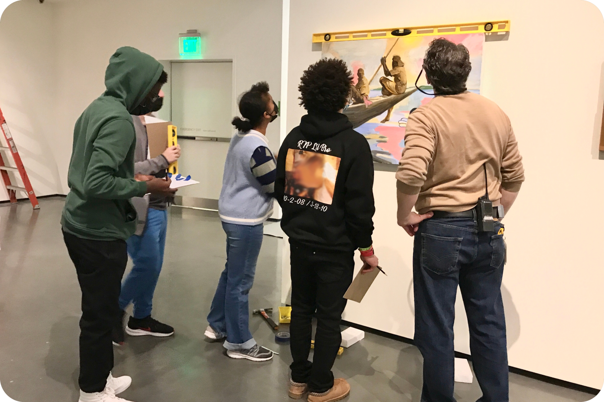 5 people standing around and looking at a painting hung on a wall. A level sits on top of the painting, and several people are holding various tools. 