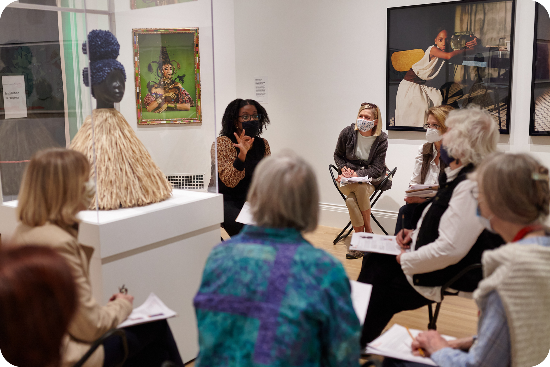 Group of people holding papers sitting around a bust sculpture encased in glass. A person in the center gestures the number three. The wall behind them features framed photographs.