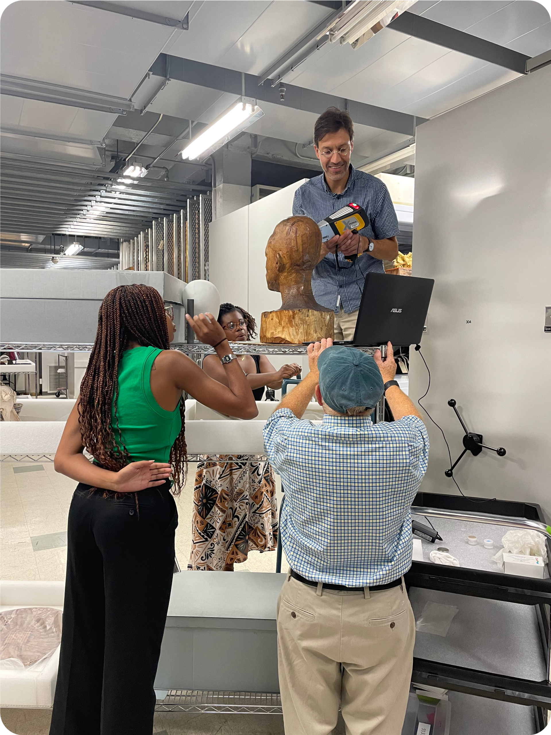 Four people around a wooden bust next to a laptop. Two people raise their arms towards the bust. Another looks sideways, while another standing on a platform holds a tool. 