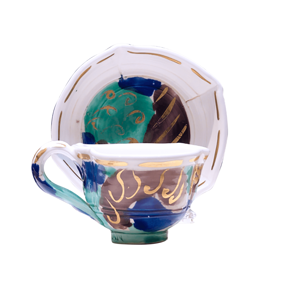 A white teacup with an upright saucer behind it. Both are covered in abstract shapes of brown, blue, and green with gold lines and squiggles.