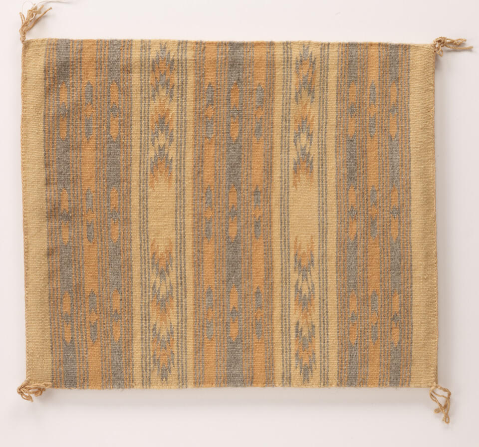 Textile with stripes. Each stripe has a subtle geometric pattern running through it. Palette is peach, tan, and dark blue. Object is square shaped, with small tassels at each corner.
