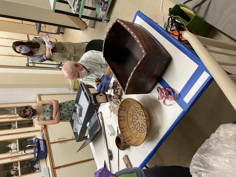 A masked person works at a table, on which are two large baskets and other objects. Two masked young people stand observing, cabinets of more objects behind them.