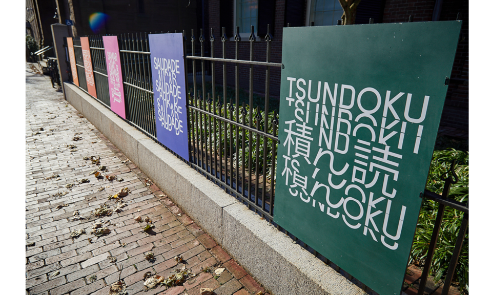 Beside a brick sidewalk, several signs are attached to a wrought-iron railing. The first sign, in green, reads “Tsundoku,” followed by stylized text.