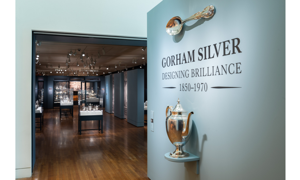Text on a light gray wall just outside an exhibition space says: “Gorham Silver: Designing Brilliance 1850-1970.” Mounted on the wall are an oversized silver coffeepot and spoon.