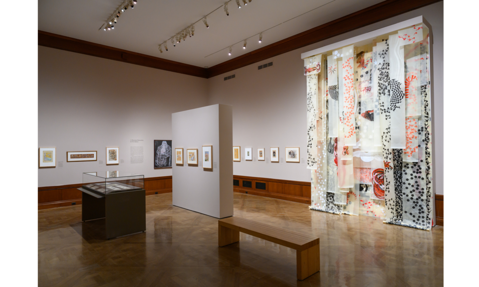 The walls of a large gallery space are hung with many works of art. At right is a colorful floor-to-ceiling installation of various decorative panels.