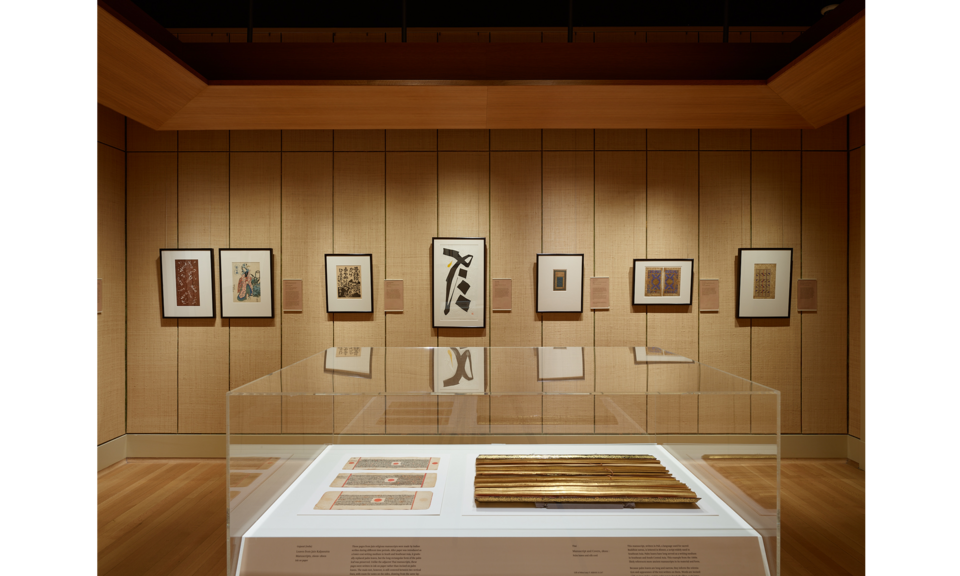 Gallery space with tan walls and peaceful lighting. Several works hang on the far wall, with additional objects in a case in the foreground.