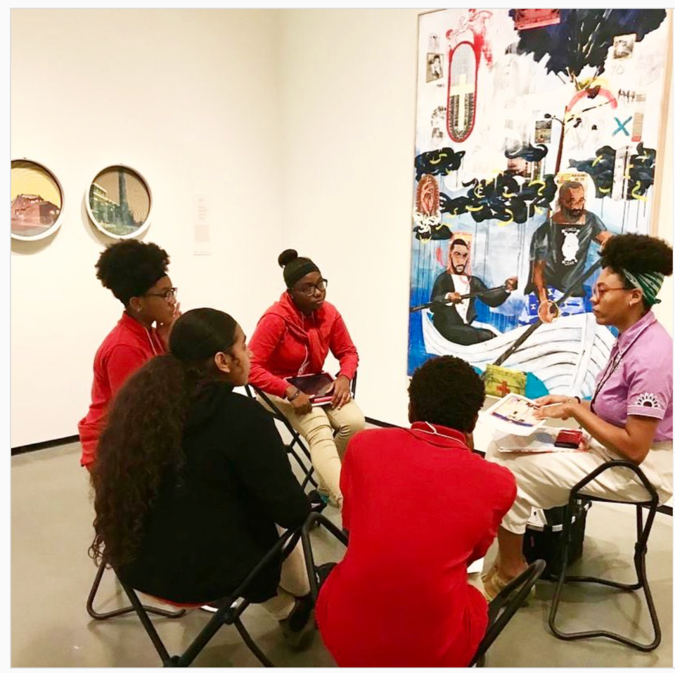 Four young dark-skinned people engaged in conversation, sitting in front of a dynamic abstract painting of two dark-skinned people rowing a boat.