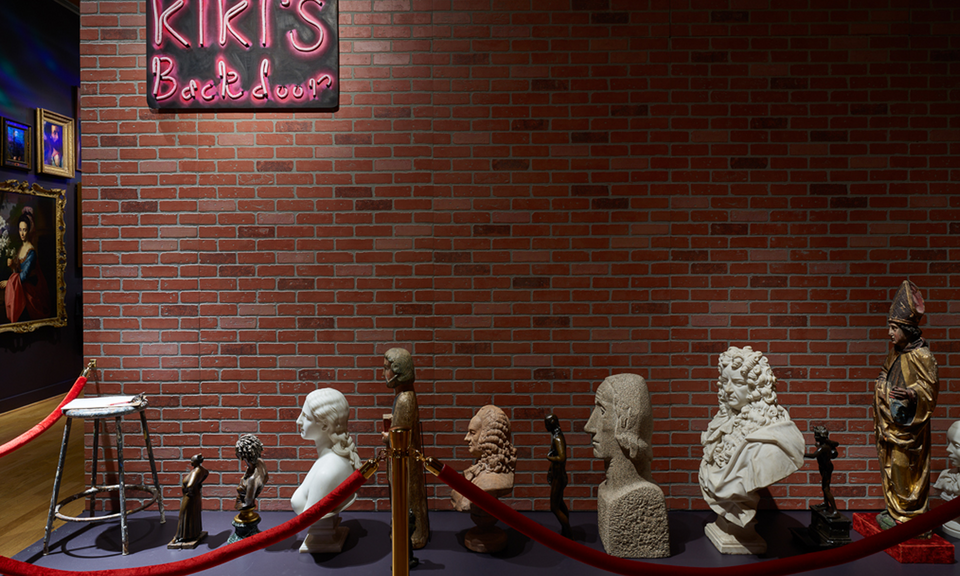 A brick wall bears a sign saying “Kiki’s Back Door.” Waiting on the ground behind a red velvet rope is a row of portrait busts and historical figurative artworks.