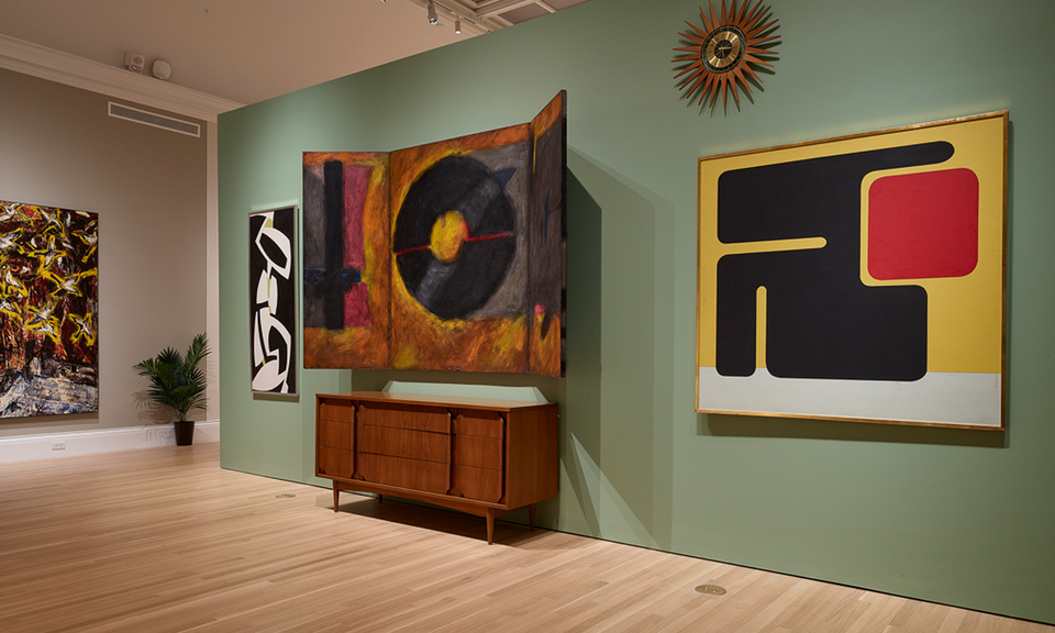 Modern paintings and furniture are displayed in a gallery space with pale green walls and wooden floors.