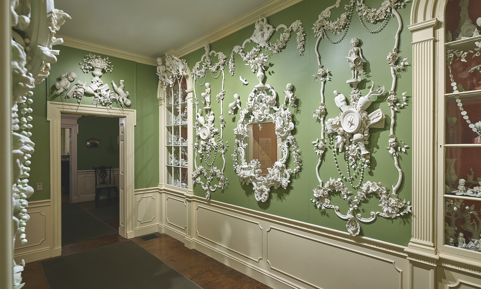 The green walls of a stately interior space featuring cream-colored woodwork are elaborately decorated with mirrors and delicate white three-dimensional figural embellishments. A built-in glass-doored cabinet is at right. 