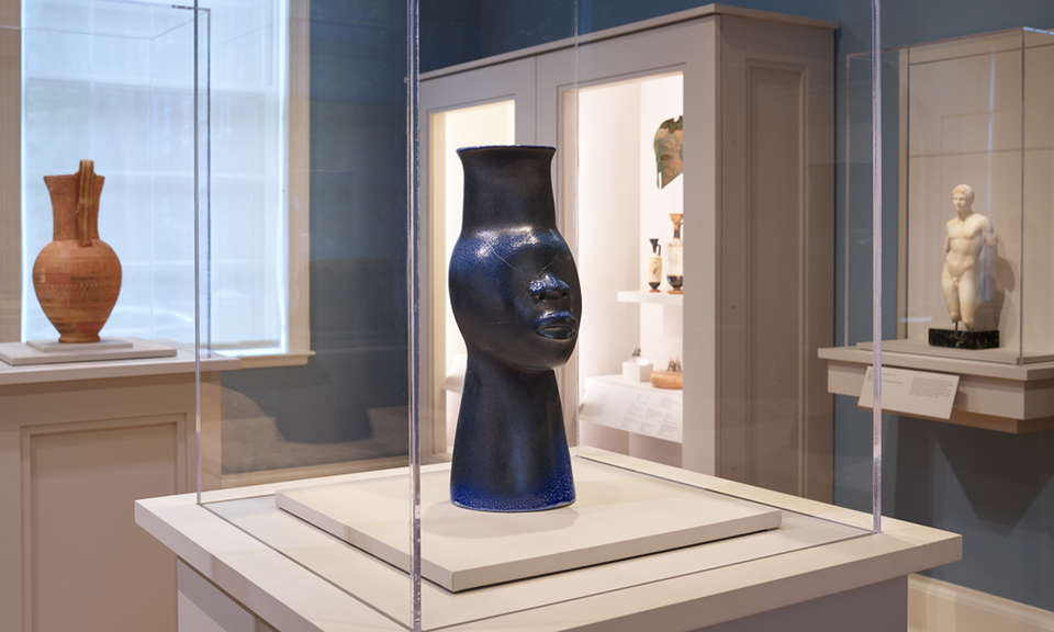 In a case in a gallery’s center is a sleek black vessel shaped like a stylized head with an elongated neck. Cases along the perimeter contain other objects.