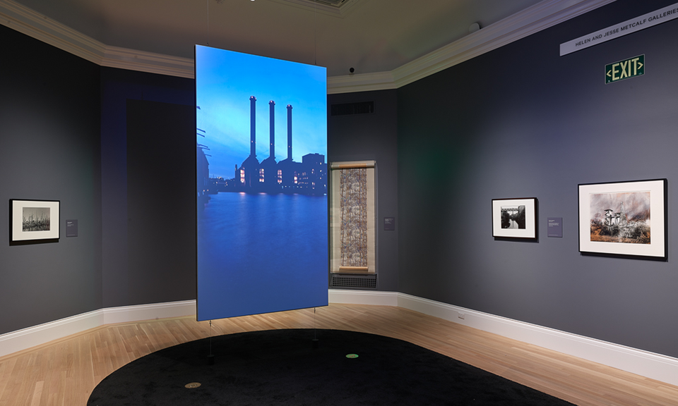 Hanging in the center of a gallery with dark-gray walls is a screen. The projected image shows an industrial building with three smokestacks. Artwork hangs on the surrounding walls.
