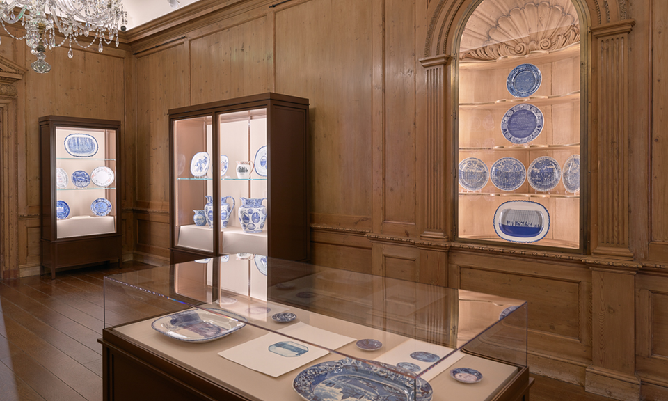 Blue and white ceramics are displayed in cases along the walls of a wood-paneled gallery. A case in the foreground contains additional works. A crystal chandelier hangs at top left.