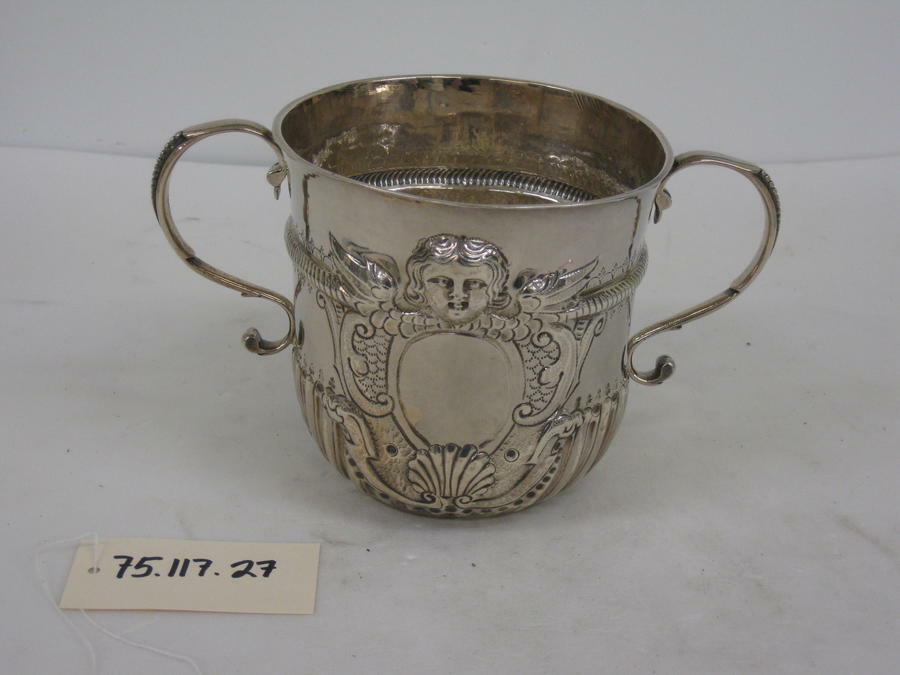 A silver cup with symmetrical handles, swirling raised designs and a small raised face on the upper half of the cup.