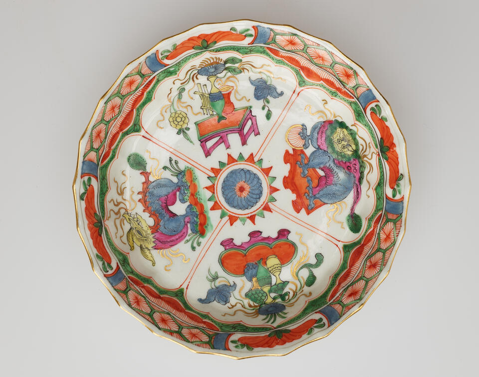 A white saucer with green, red, blue, pink, yellow, and gilded decorations; separated into four main vignites, two animals and two tables and vessels containing floral elements.
