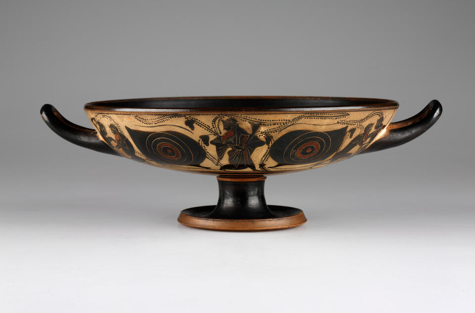 A wide, shallow, footed vessel with two handles on either side, curving upwards. The body is black and tan colored with an abstract design. 