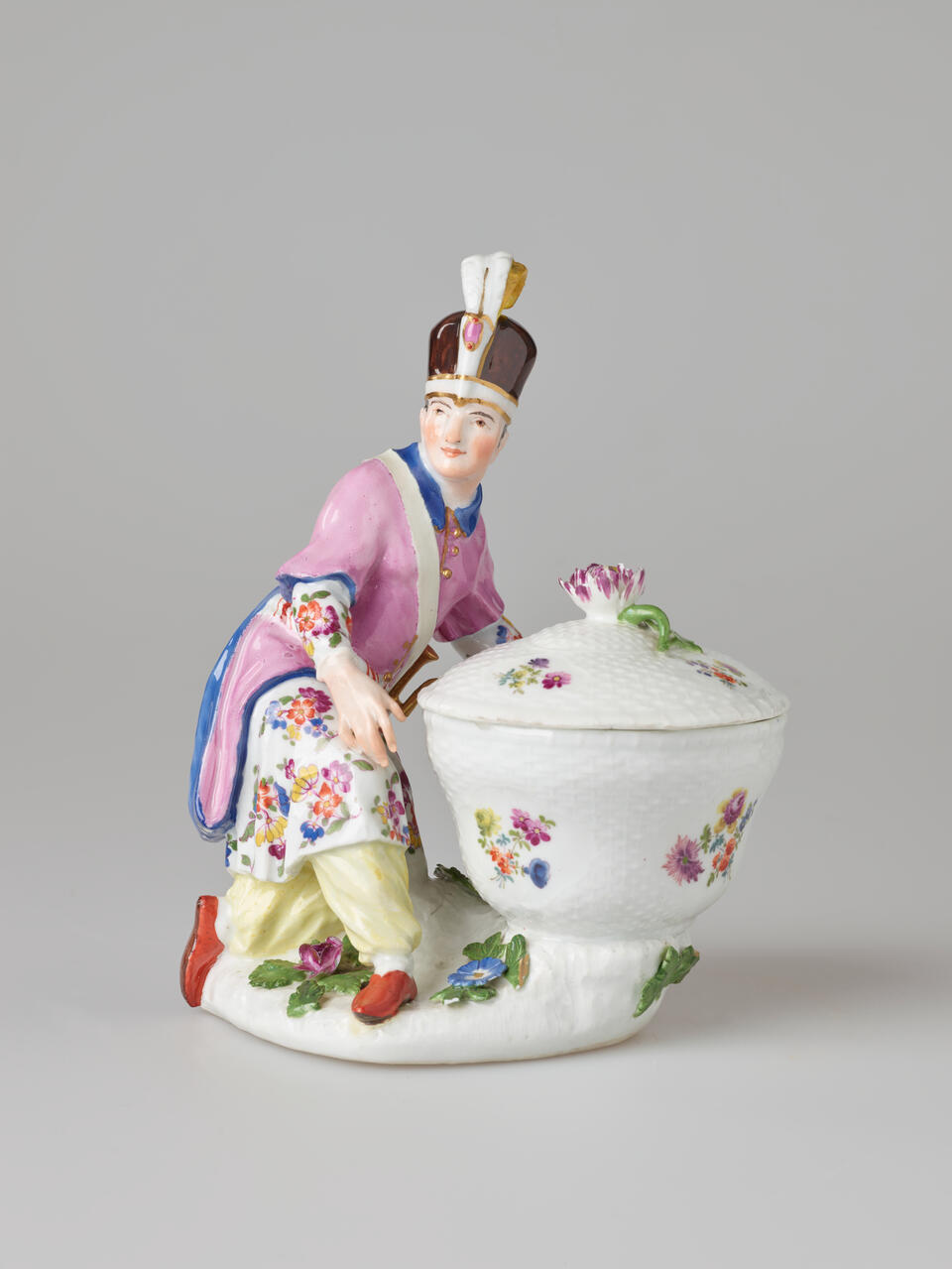 A sculptural figure kneeling next to a basket in bright elaborate clothing with a hat with a feather. There are floral decorations on the figure’s clothing and basket.