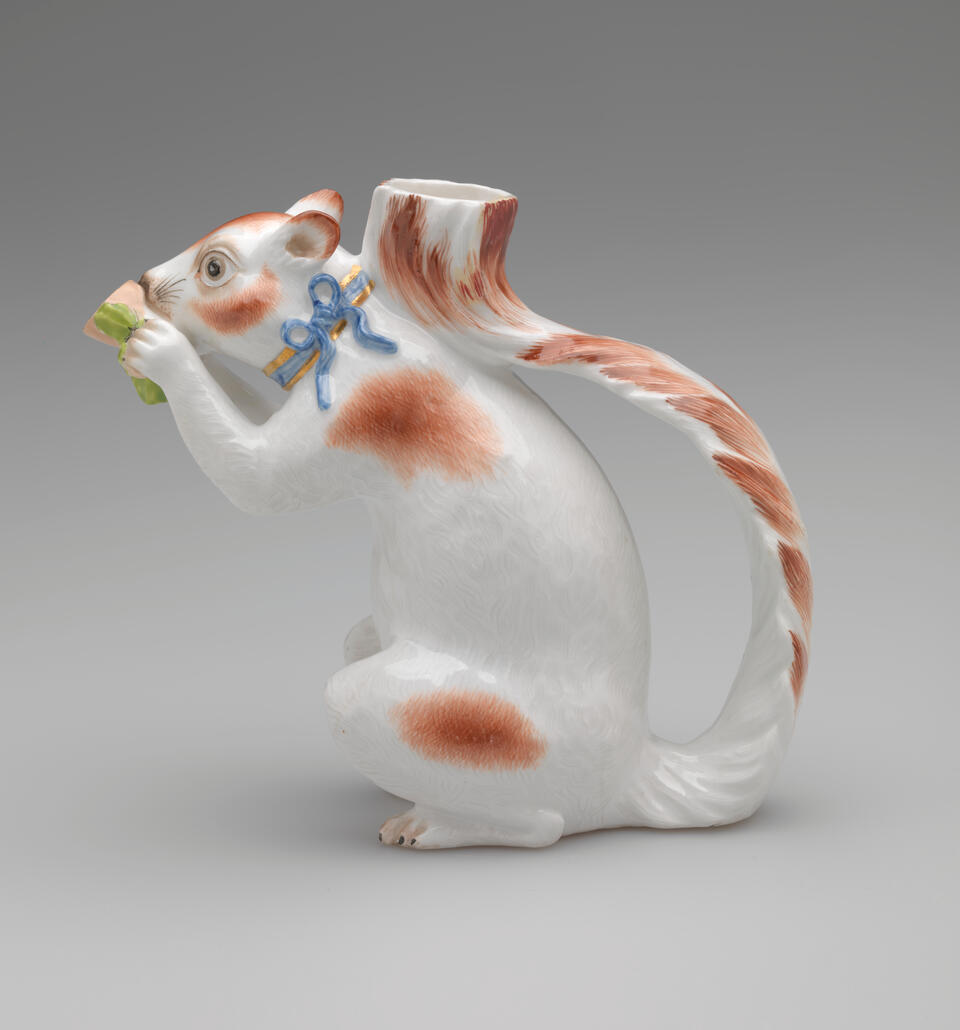 Vessel in the shape of a white squirrel with brown patches and a blue ribbon arounds its neck. It holds a brown nut with green leaves up to its face.