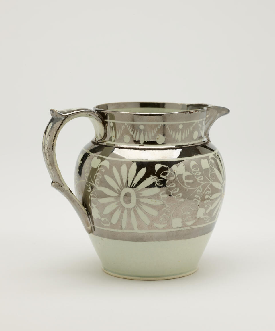 A silver ceramic jug with a decorative handle, there are decorative floral motifs on the body of the vessel.