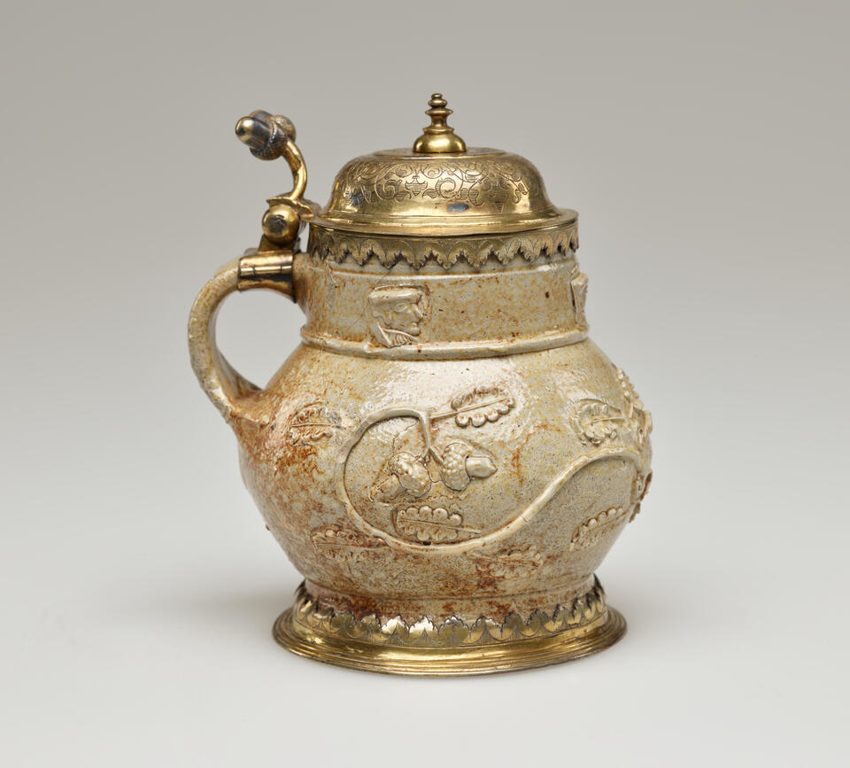 An unglazed stoneware mug with tan to copper colors and floral decoration. The foot goes into a gilded metal decoration, similarly to the handle where the hinged lid is attached.