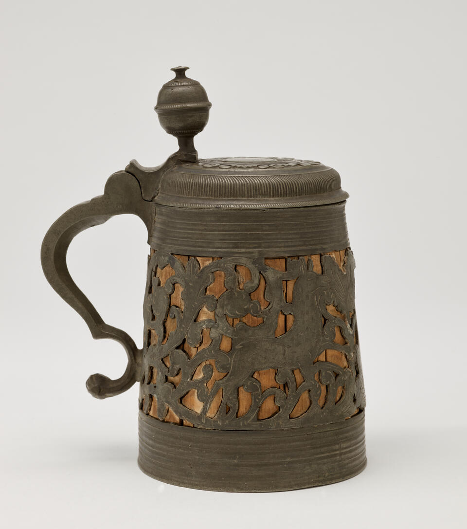 A wood and pewter mug that has a hinged lid and handle. The pewter is cut out and shows the wood on the interior. 