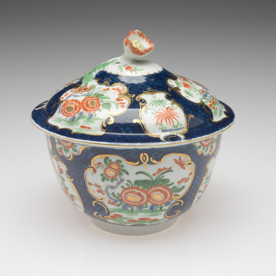 A lidded sugar bowl with floral decorations in gilded vignettes, surrounded by dark blue glaze, the finial on the lid is a sculptural red, white, and yellow flower. 