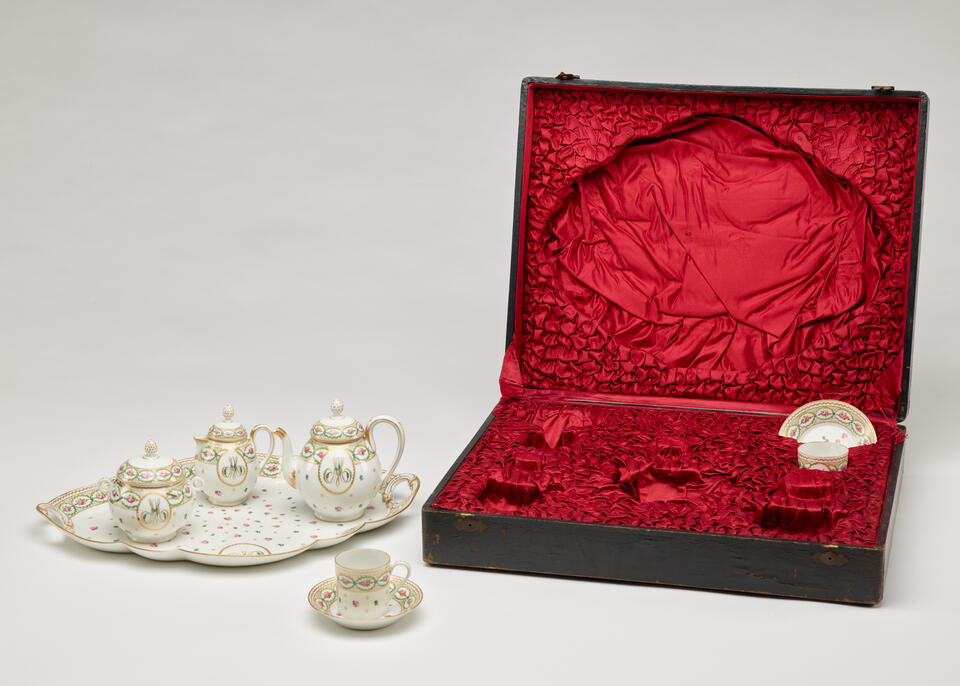 A white, pink, green, and gilded tea set with floral decorations and consists of four vessels and a tray. The traveling case with a black exterior and red fabric interior. 