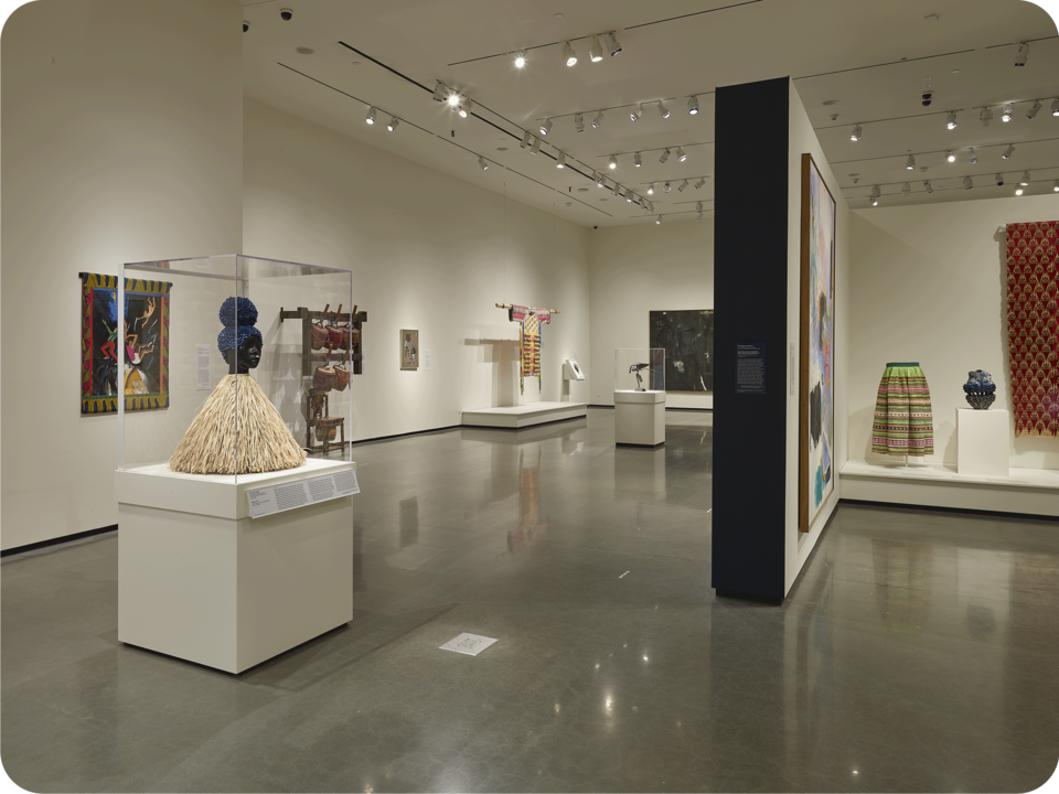 Installation view of an exhibit. A partial wall with a black painted side extends towards the viewer. Visible are colorful textile works, paintings, ceramics, and figurative sculptures. 
