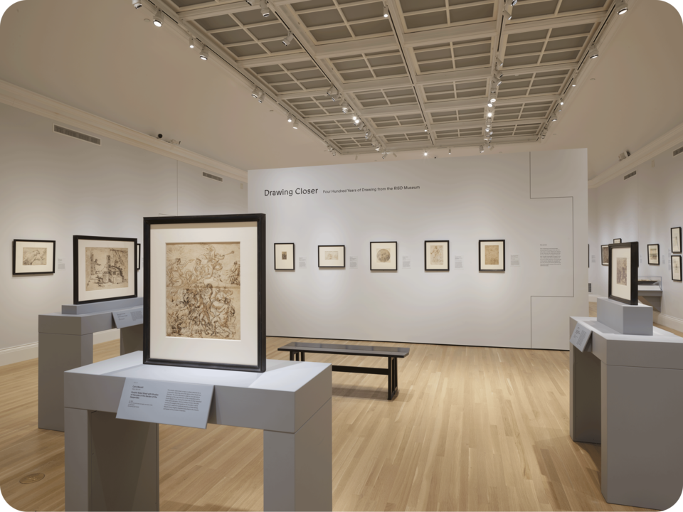 Installation view of an exhibit, with several drawings propped on gray tables, and other drawings hung on the wall. A central divider reads “Drawing Closer”. In front of it is a bench. 