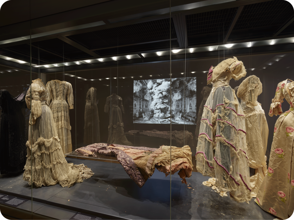 View of several ornate cream-colored dresses on mannequins encased in glass. A table in the center holds another dress. In the background is a projection of a grand ornate space. 