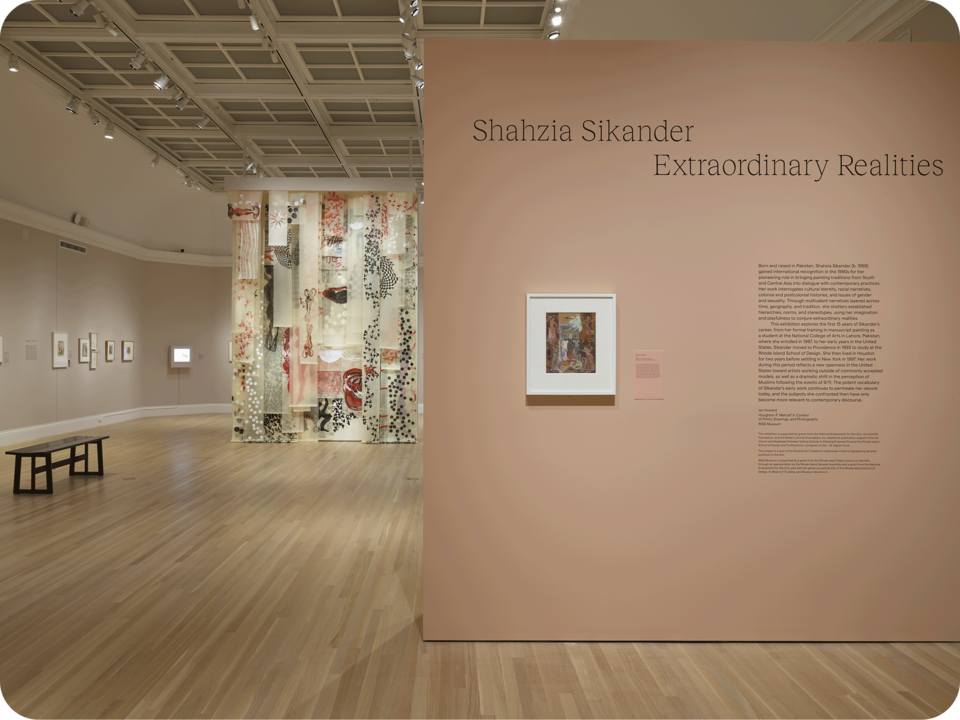 Exhibition documentation. A partial wall in the foreground features artwork and exhibition information, reading “Shahzia Sikander Extraordinary Realities”. Behind it are more artwork on walls, and a larger work suspended from the ceiling. 