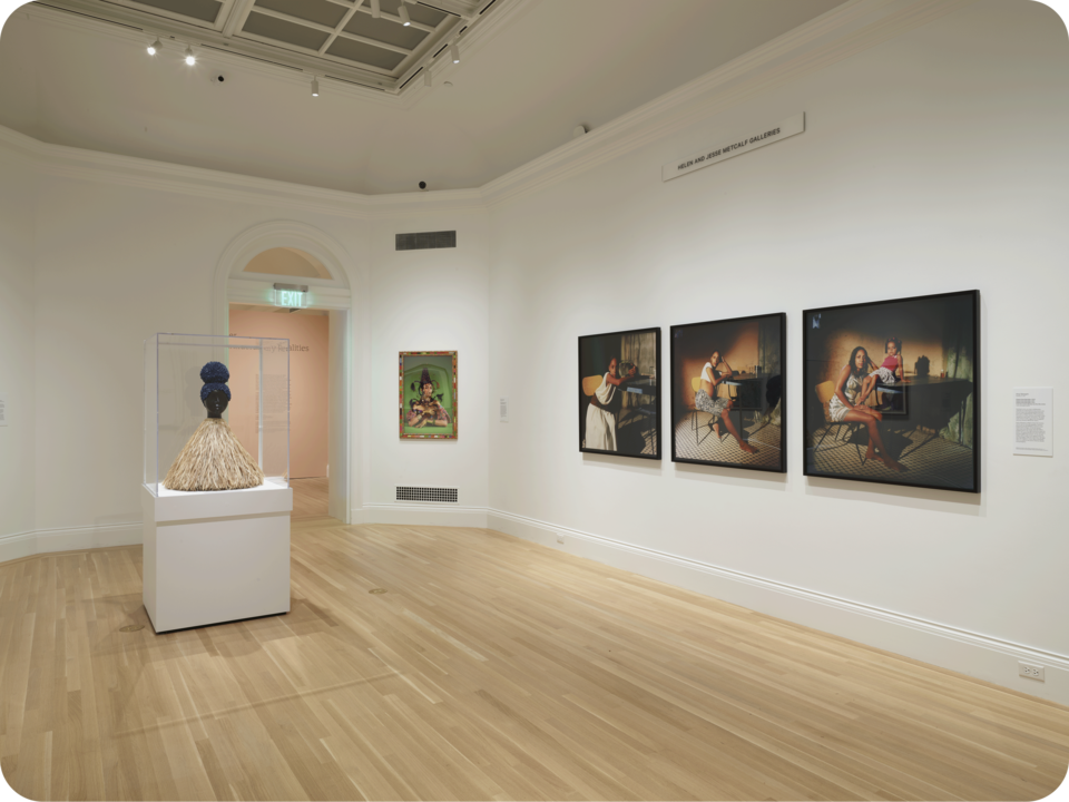 Exhibition documentation. 3 photographs in a similar style are hung on one wall, with another photo on the adjacent wall. A bust in a glass case occupies the space’s center.