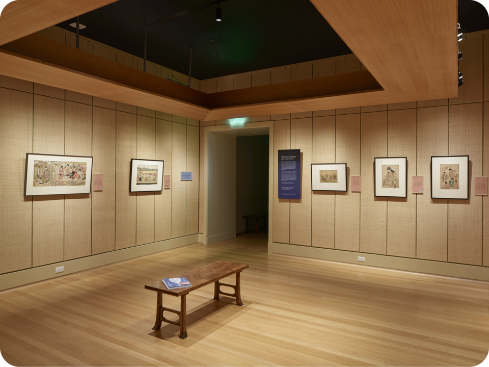 Exhibit documentation. Two framed prints hang on one wall, with three other prints and exhibition text on another wall, separated by a doorway. A wooden bench occupies the space’s center. 