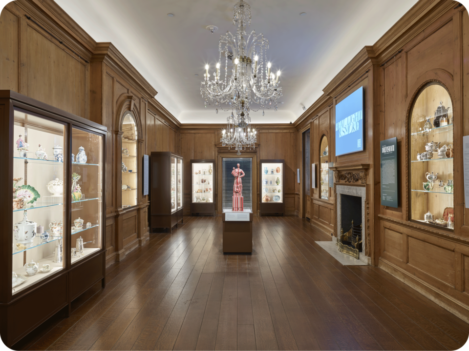 Exhibit documentation. Cases containing ornate ceramics and metalwork line the three walls of the space. A striped statue stands in the space’s center below a large chandelier. 