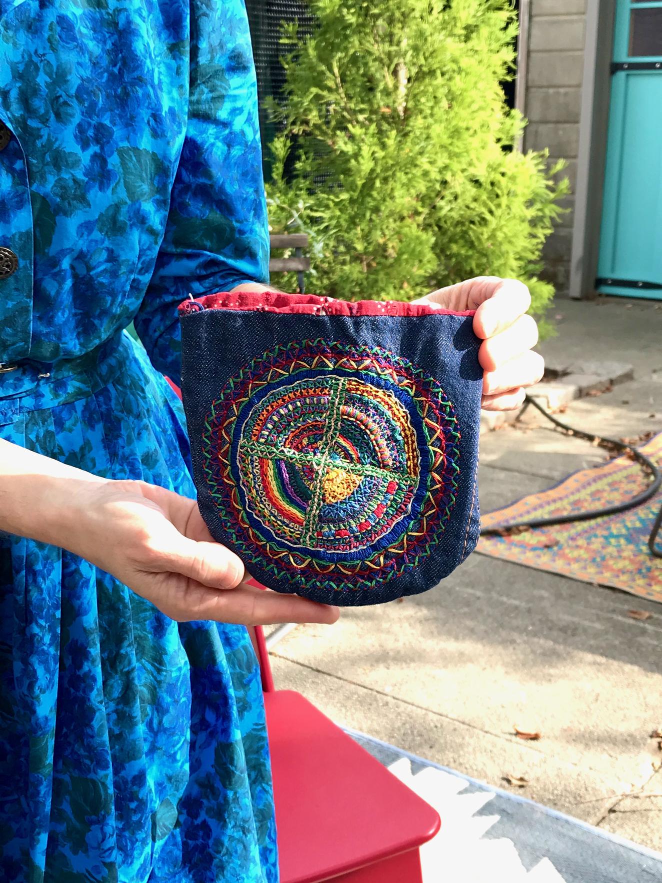 Color photograph, a close-up of a small denim purse with rainbow embroidery in a radial pattern. The photograph is outside, and the purse is held by a white woman in a blue floral dress. We only see her hands and torso.